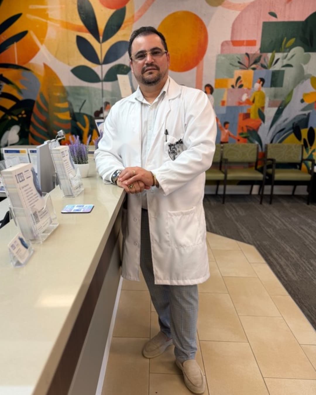 Dr. Hendry Pascual standing at the reception counter at PassQual Health, smiling in his white coat, with the community mural behind him