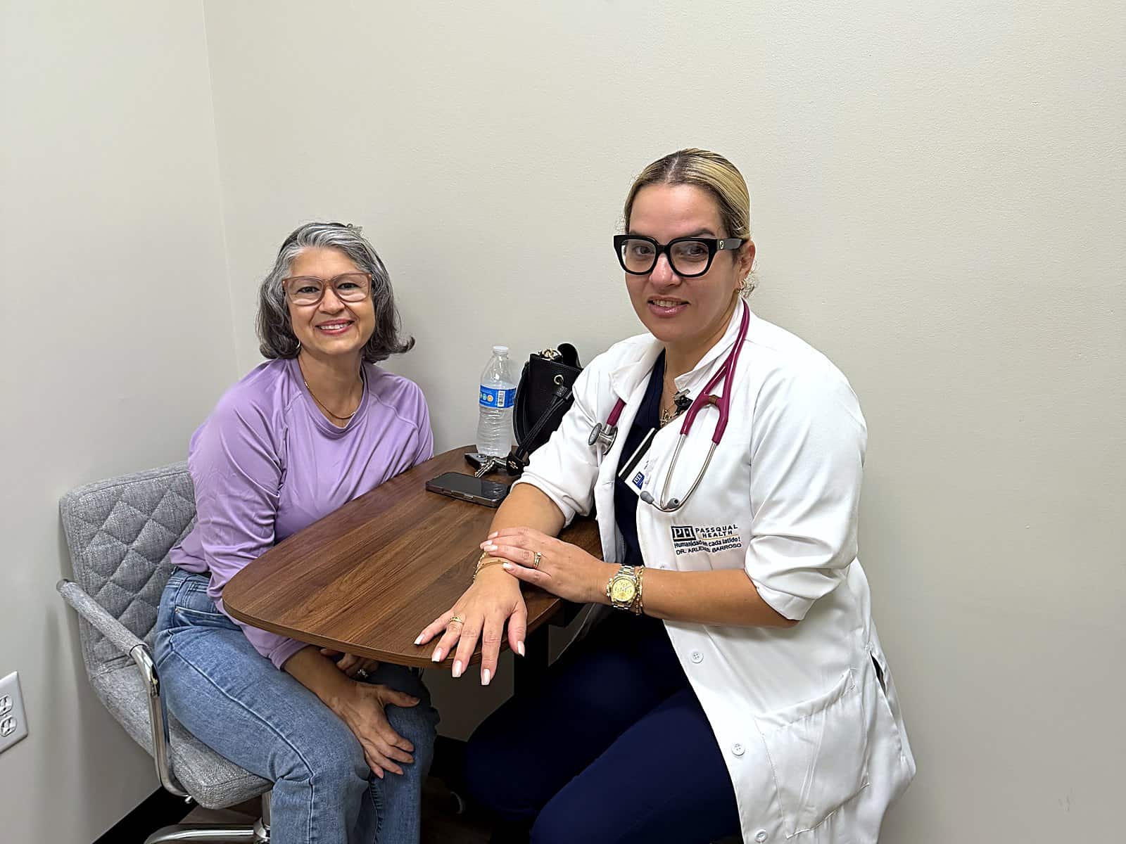 A PassQual Health physician in a white coat consulting with a patient at a table inside an exam room