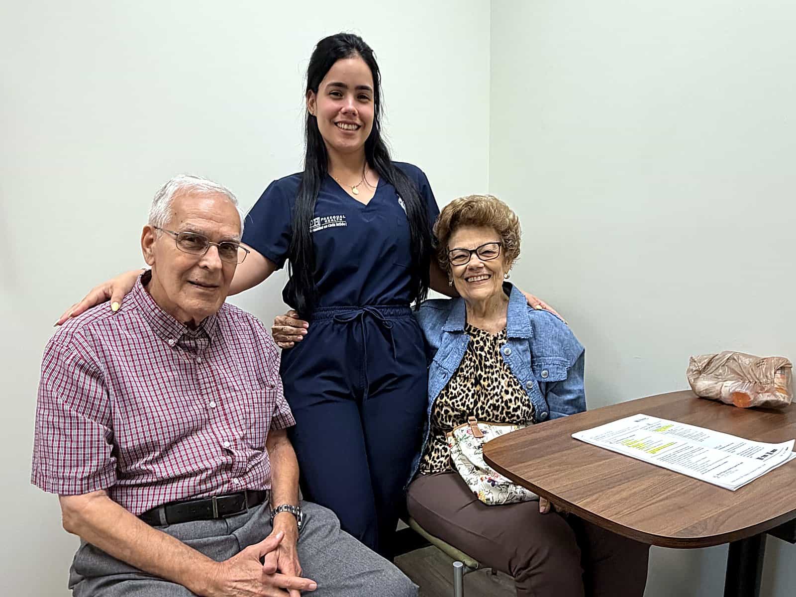 A PassQual Health team member standing with an older couple, arms around both, in an exam room