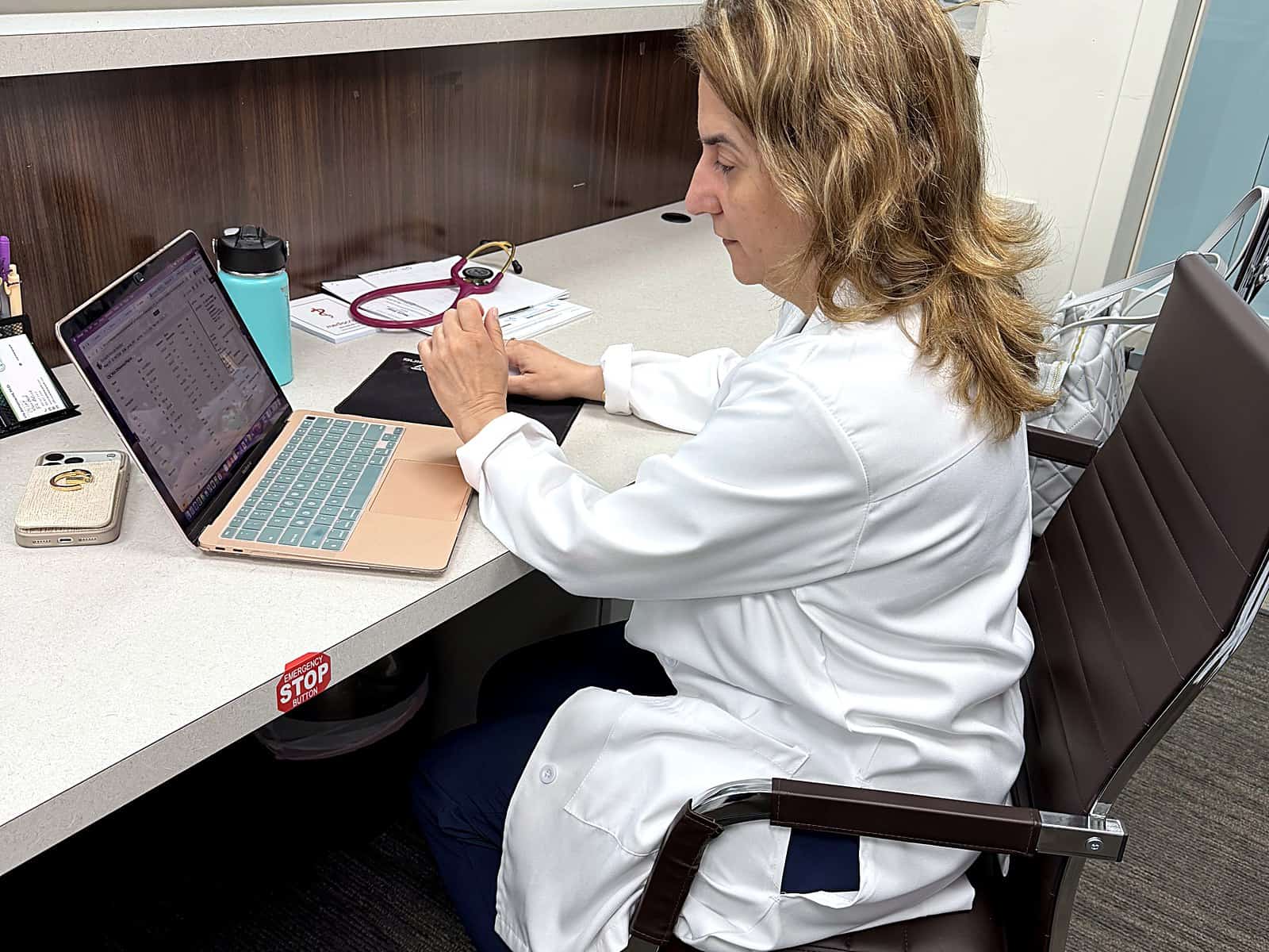 A PassQual Health physician in a white coat working at her desk with a laptop, the kind of setup used for virtual visits