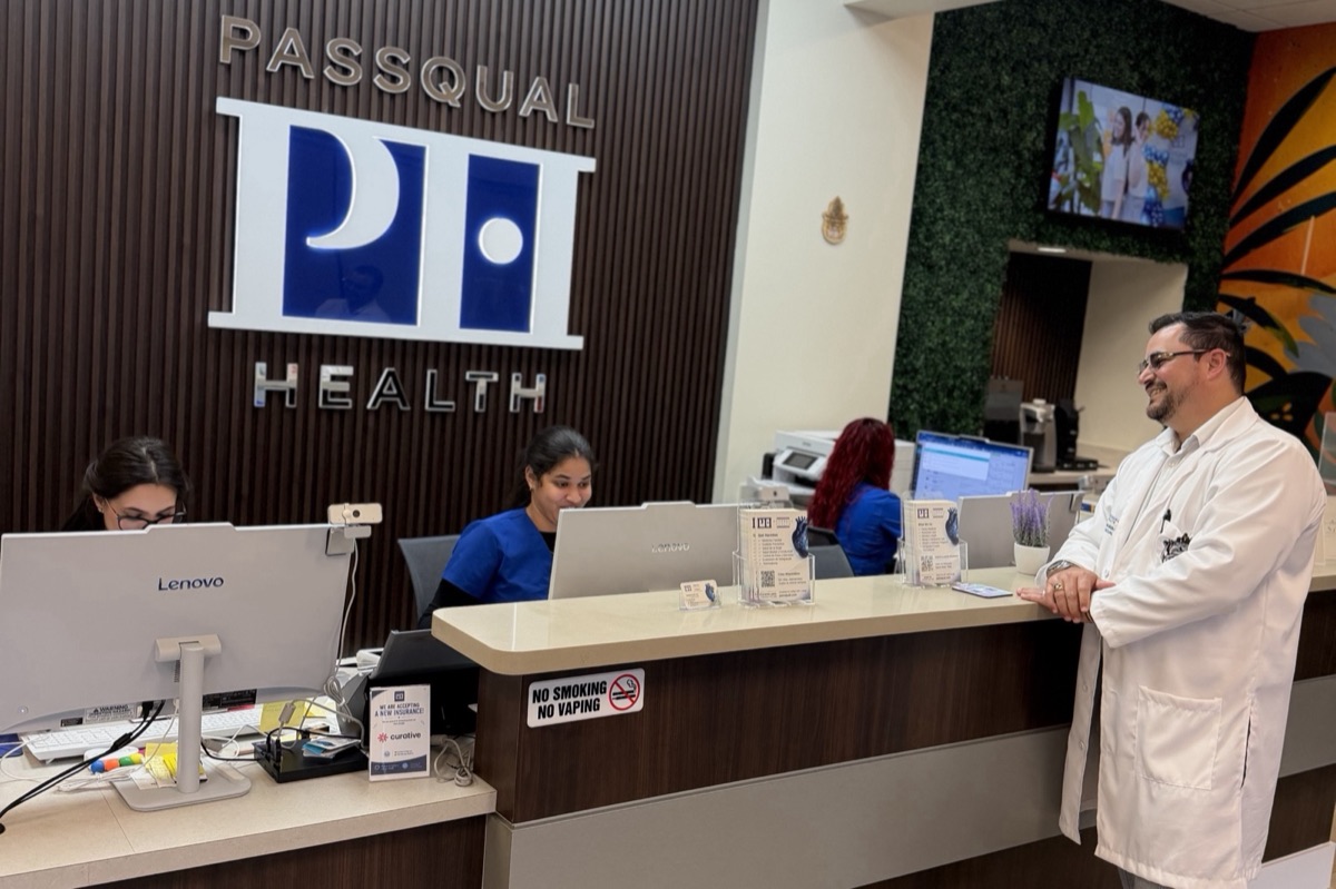 PassQual Health reception area in Miami Gardens with the dimensional metal "PASSQUAL PH HEALTH" wall sign, three staff members at work, and Dr. Pascual in his white coat smiling toward his team, with the community mural and green wall behind them